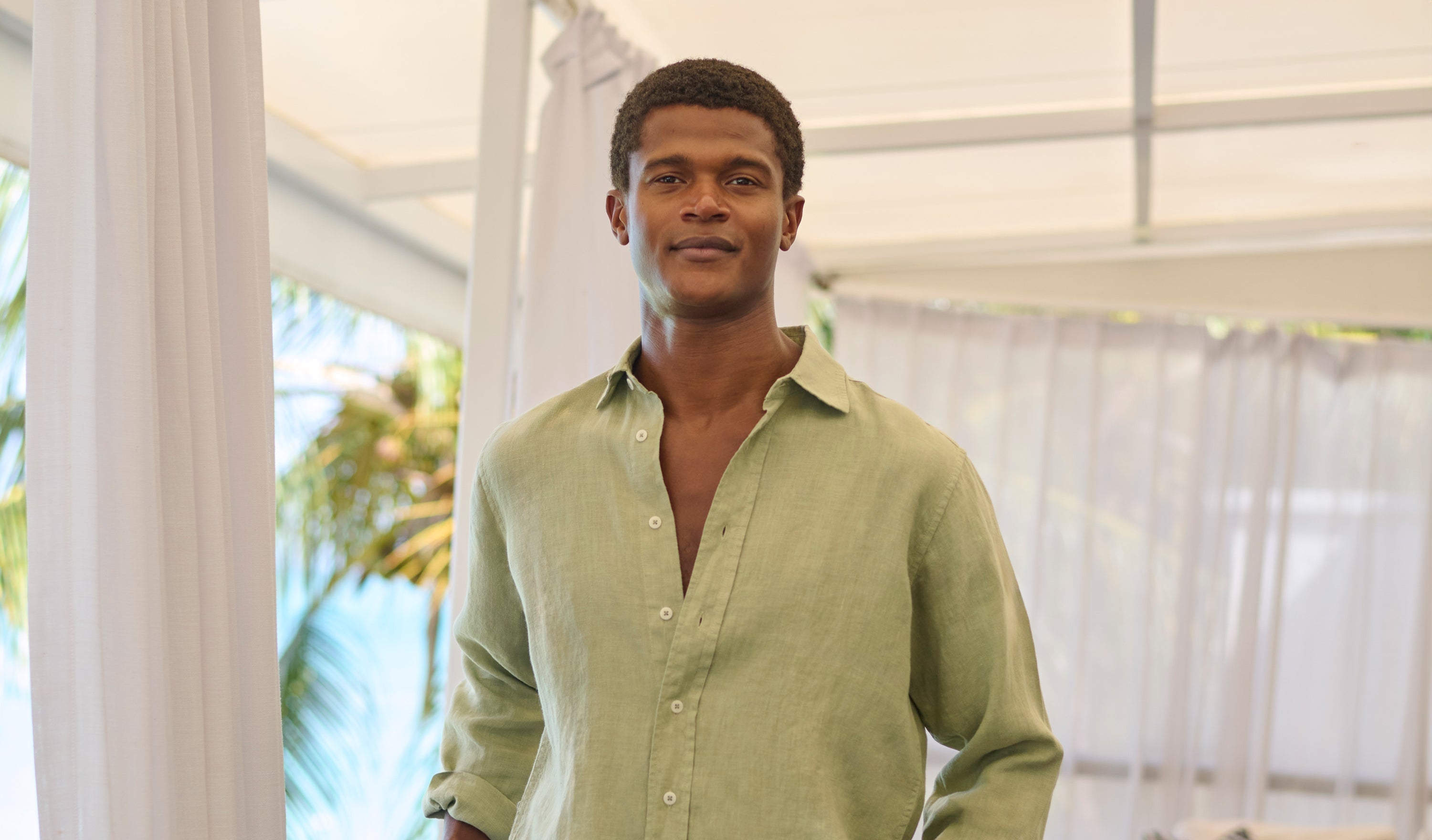 Man wears green linen shirt in an open air room with palm trees in the background