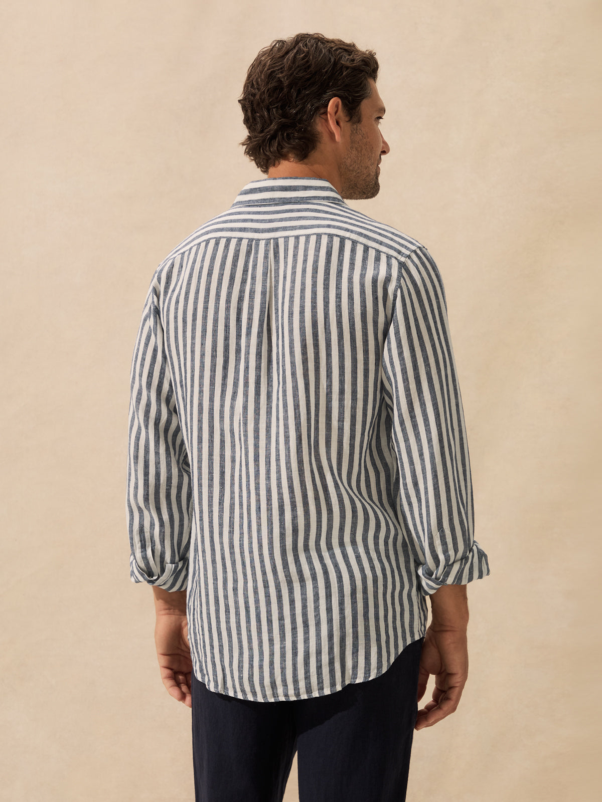 A man shown from the back wearing a long-sleeve button-up shirt with vertical navy and off-white stripes, paired with dark trousers, against a neutral beige background.