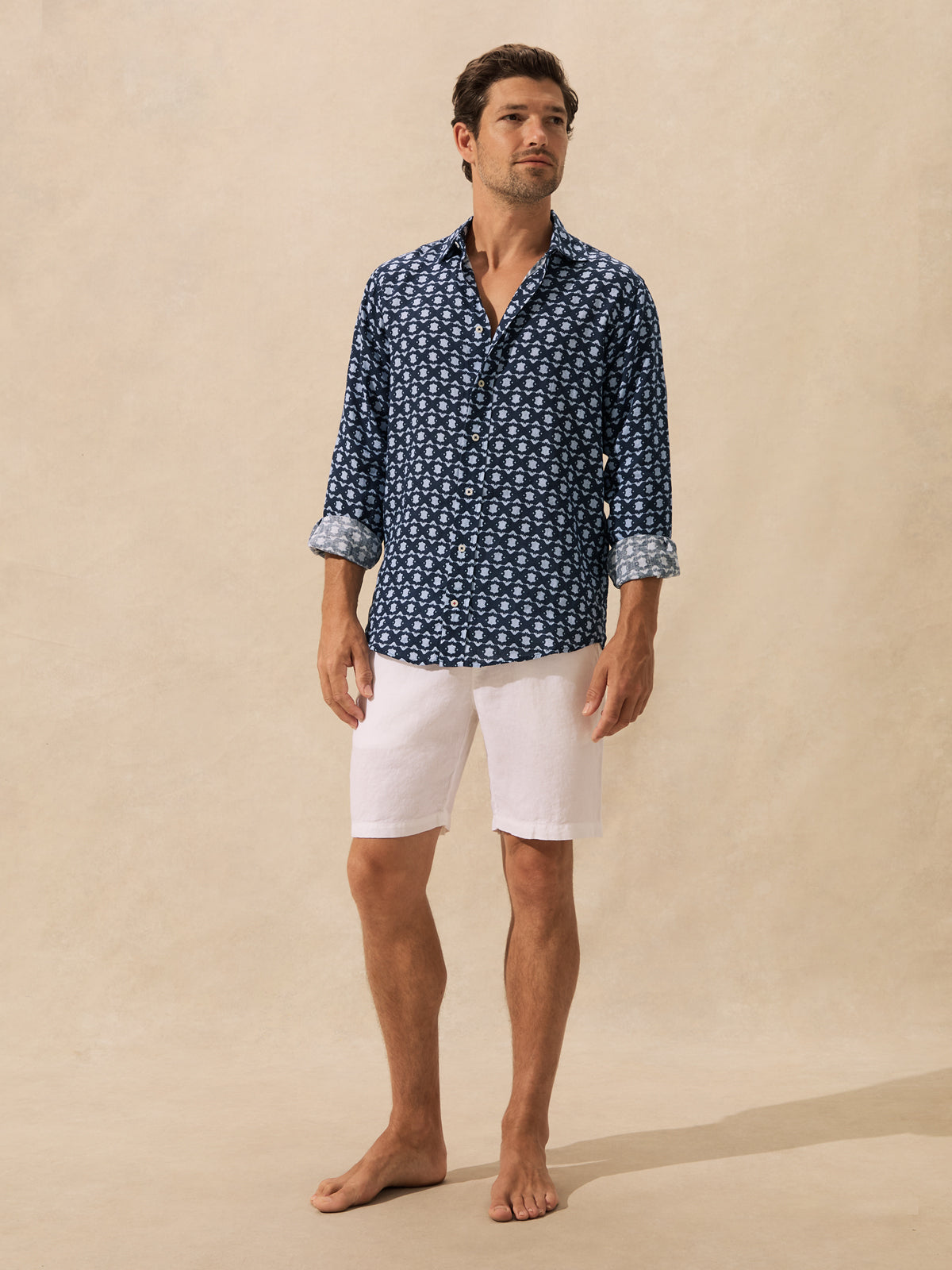 A man stands barefoot against a warm beige studio backdrop, wearing a navy blue long-sleeve button-up shirt with a small white allover pattern and white shorts.