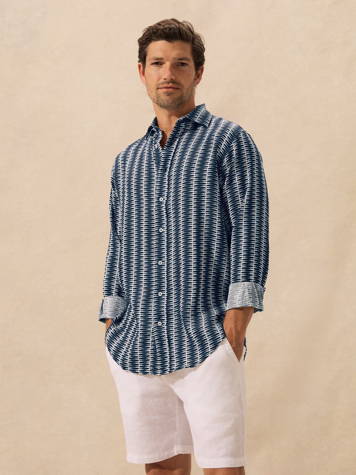 A man wearing a long-sleeve button-up shirt with a blue and white geometric pattern, standing against a textured beige background.