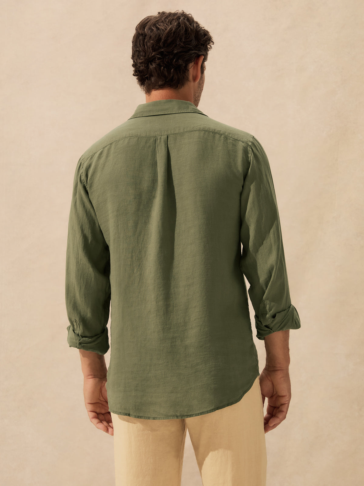 A man viewed from behind wearing an olive green long-sleeve linen shirt and beige trousers, standing against a textured beige background.