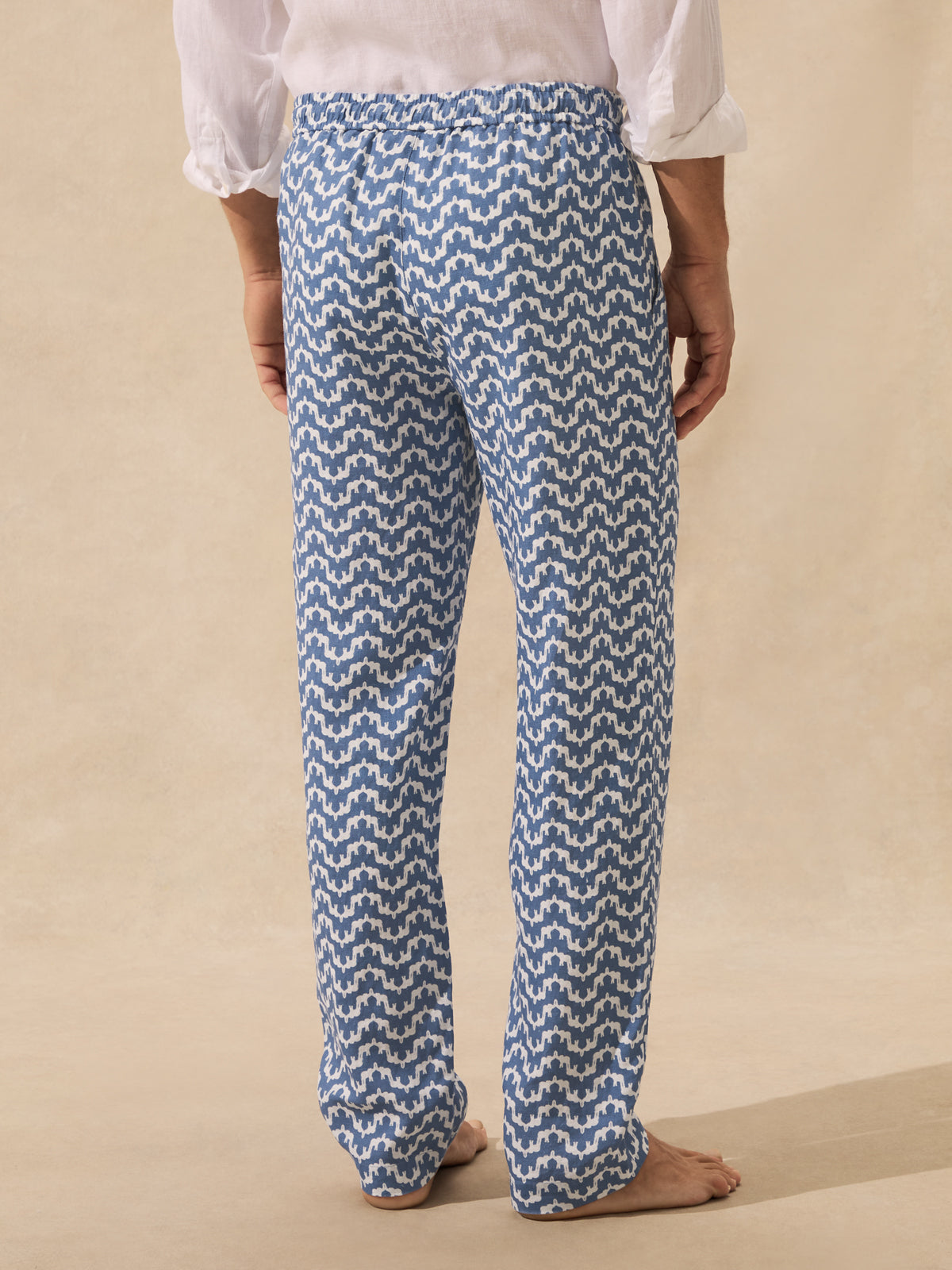A person shown from the back wearing loose blue and white patterned pants, standing barefoot against a warm beige studio background.