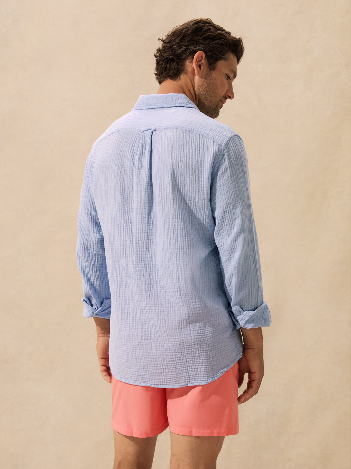 A man shown from the back wearing a light blue and white gingham long-sleeve button-down shirt and coral pink shorts, standing against a textured beige background.