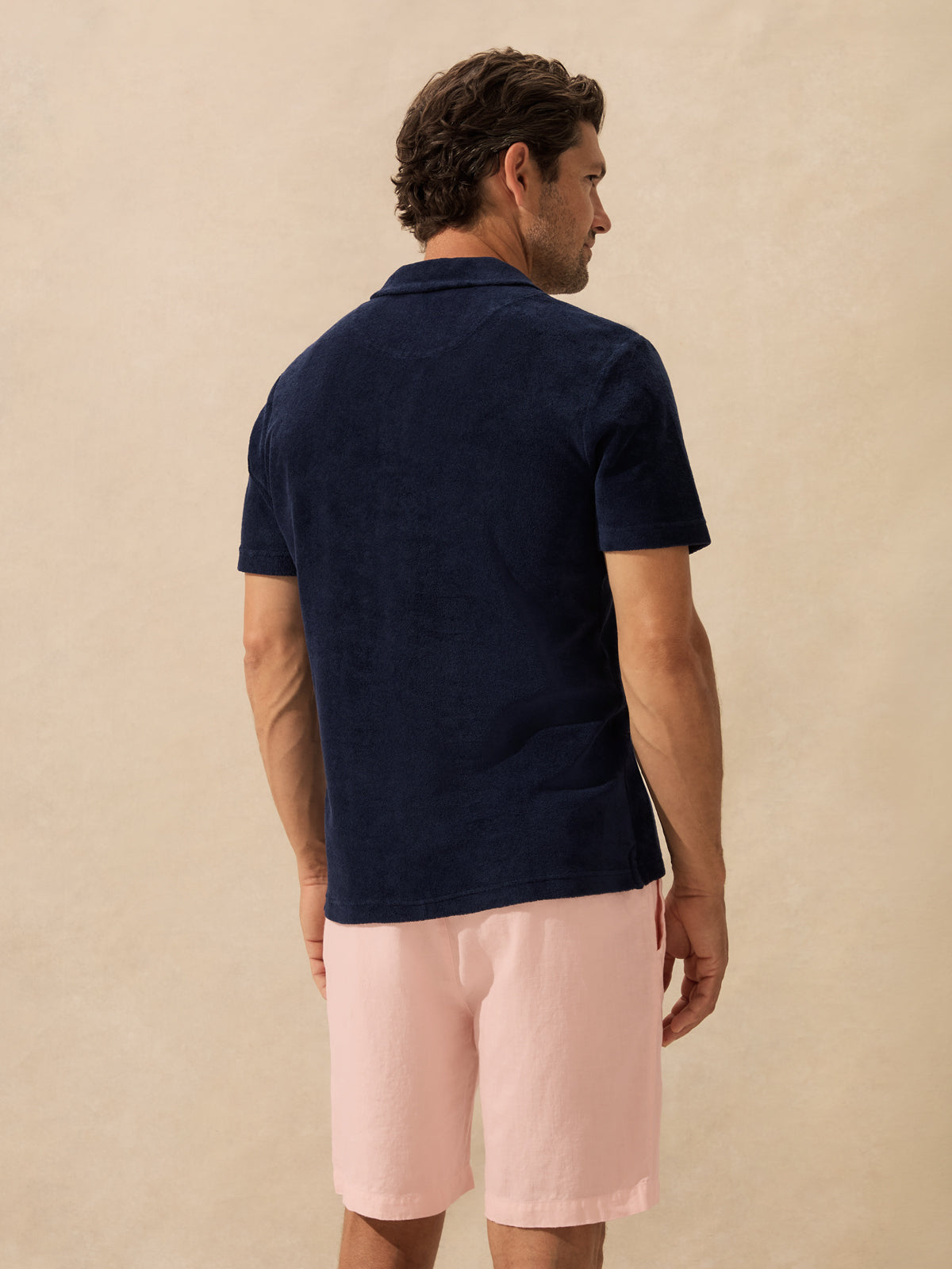 A man shown from the back wearing a dark navy short-sleeve polo shirt and light pink shorts against a textured beige studio background.