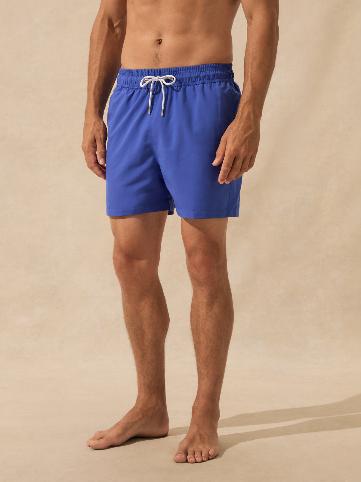 A man wearing bright royal blue swim shorts with a white drawstring, standing barefoot against a warm beige textured studio backdrop.