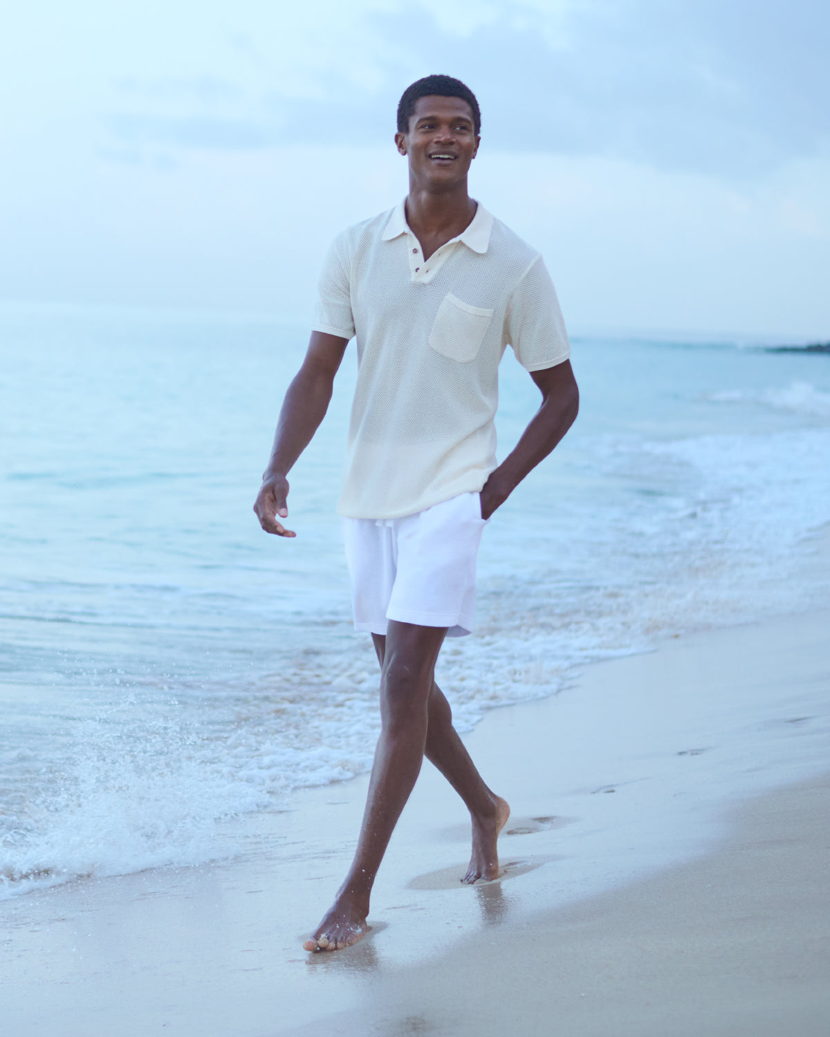 Man walking on a beach wearing a knitted polo shirt and white terry towel shorts.