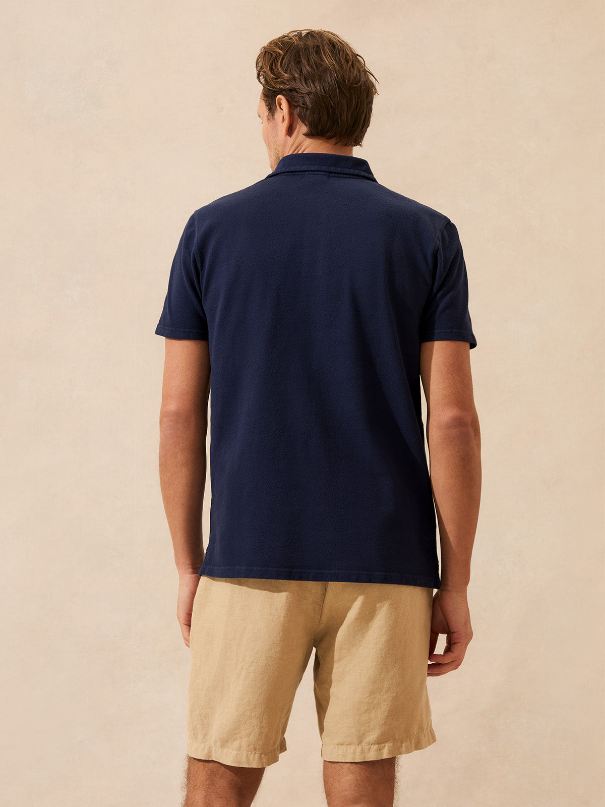 Back view of a man wearing a navy polo shirt and beige shorts on a beige background