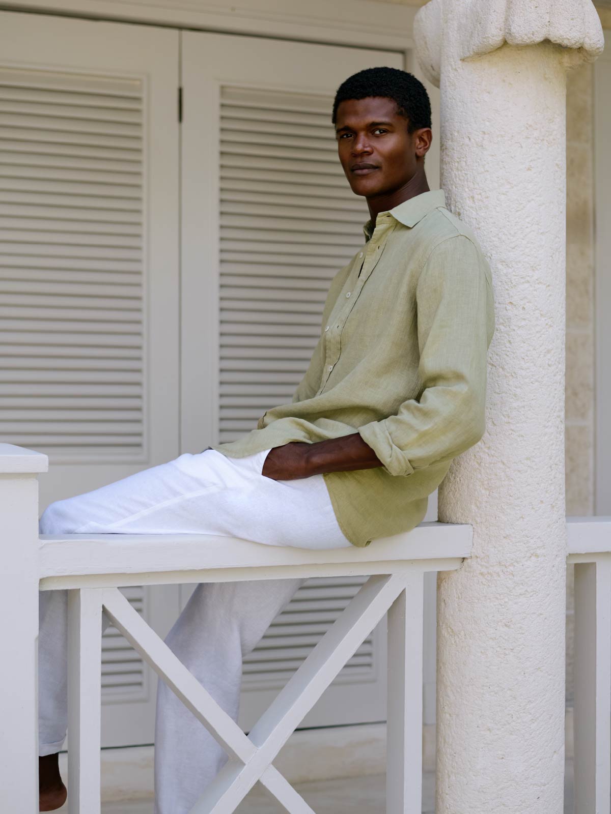 Man sitting on a white fence wearing a light green linen shirt and white linen pants, leaning against a white column.