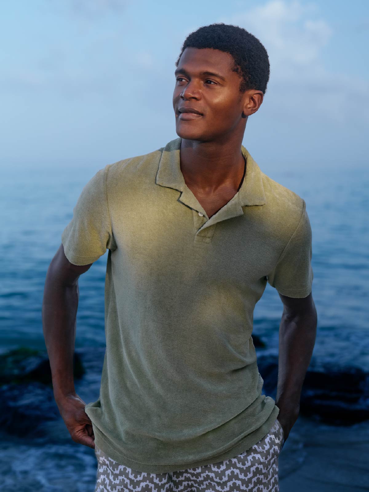 Man wearing a green terry cotton polo shirt and patterned swim shorts standing by the water.
