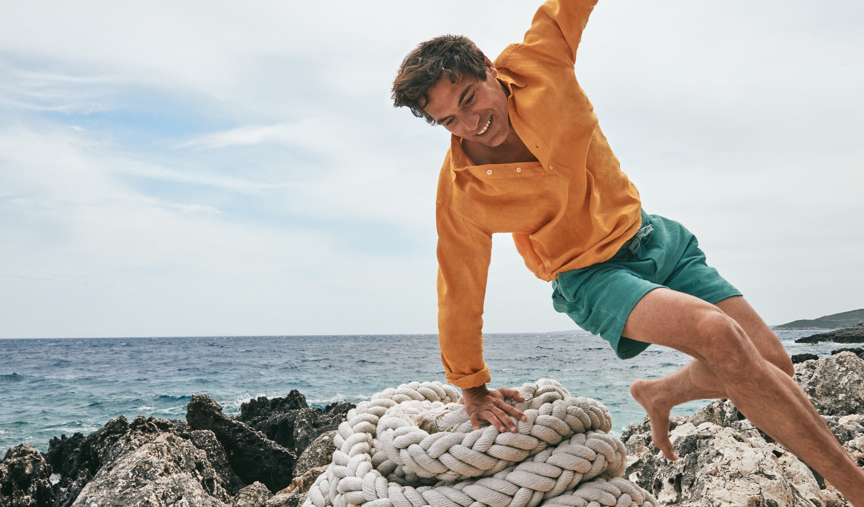 Person in yellow linen shirt and green terry shorts playing with a rope on a rocky beach.