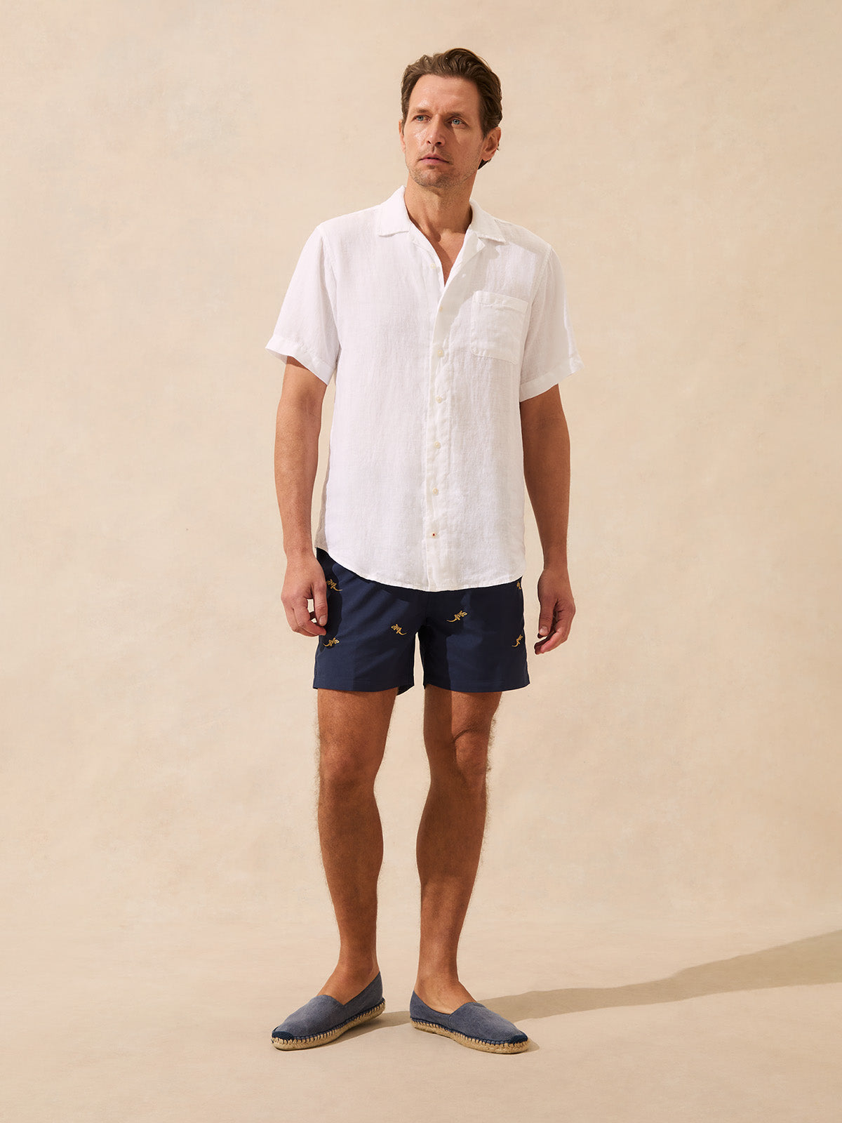 Man wearing a white short sleeved linen shirt and navy embroidered swim shorts on a beige background