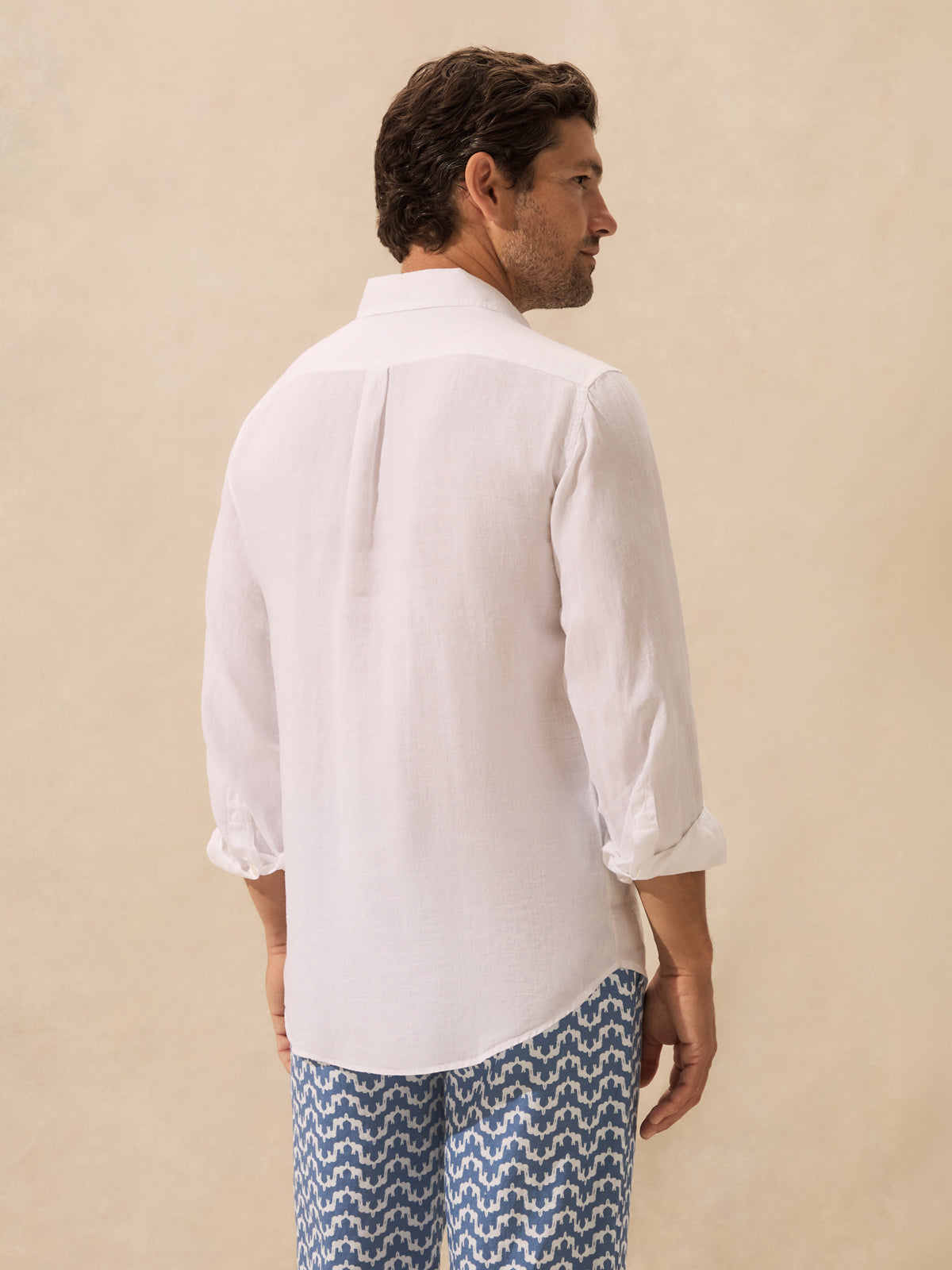 A man shown from the back wearing a white long-sleeve button-up shirt with rolled cuffs and blue patterned trousers, standing against a plain beige studio background.