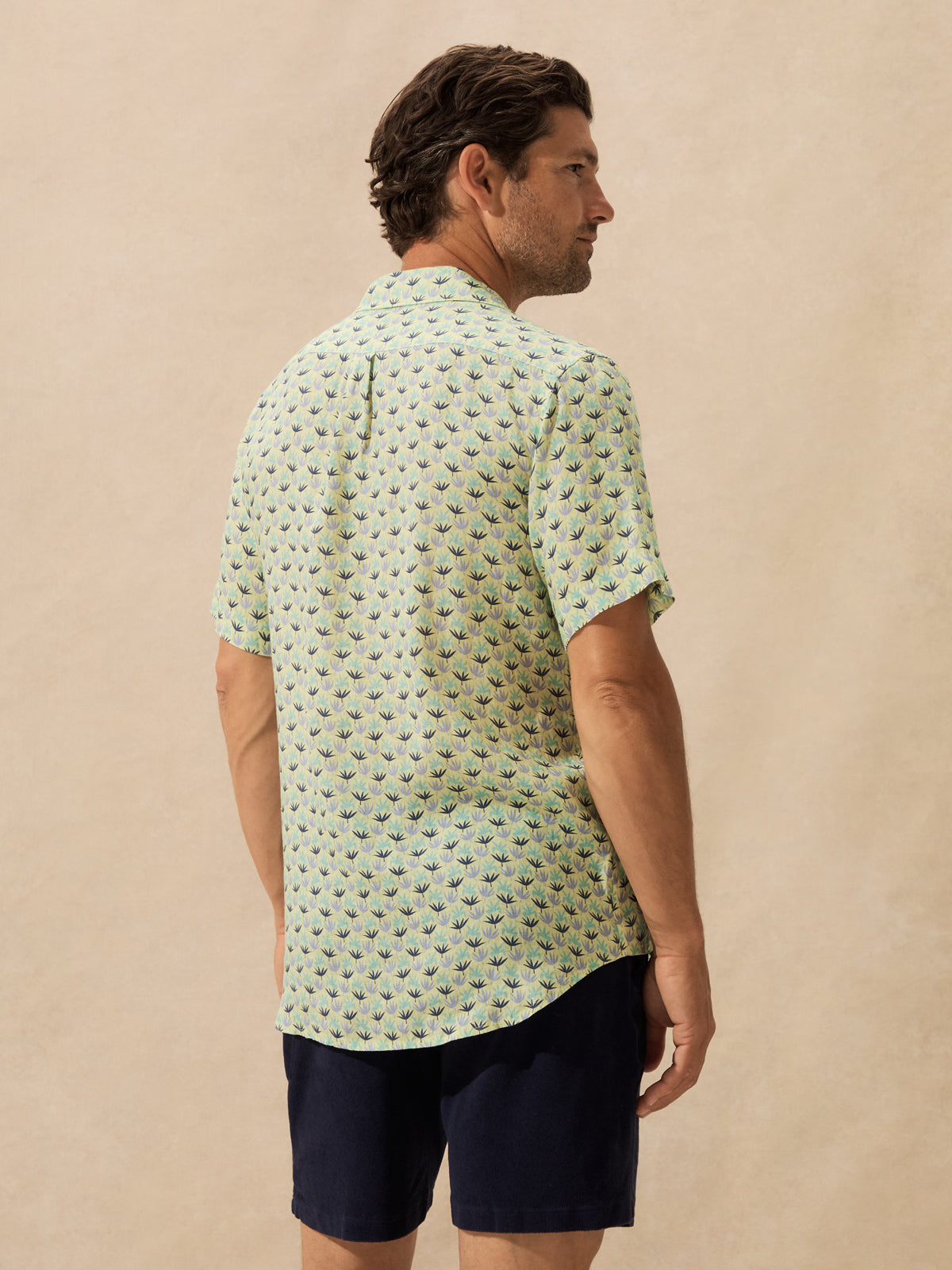 A man wearing a light green short-sleeve button-up shirt with a small repeating tropical pattern, shown from the back. 