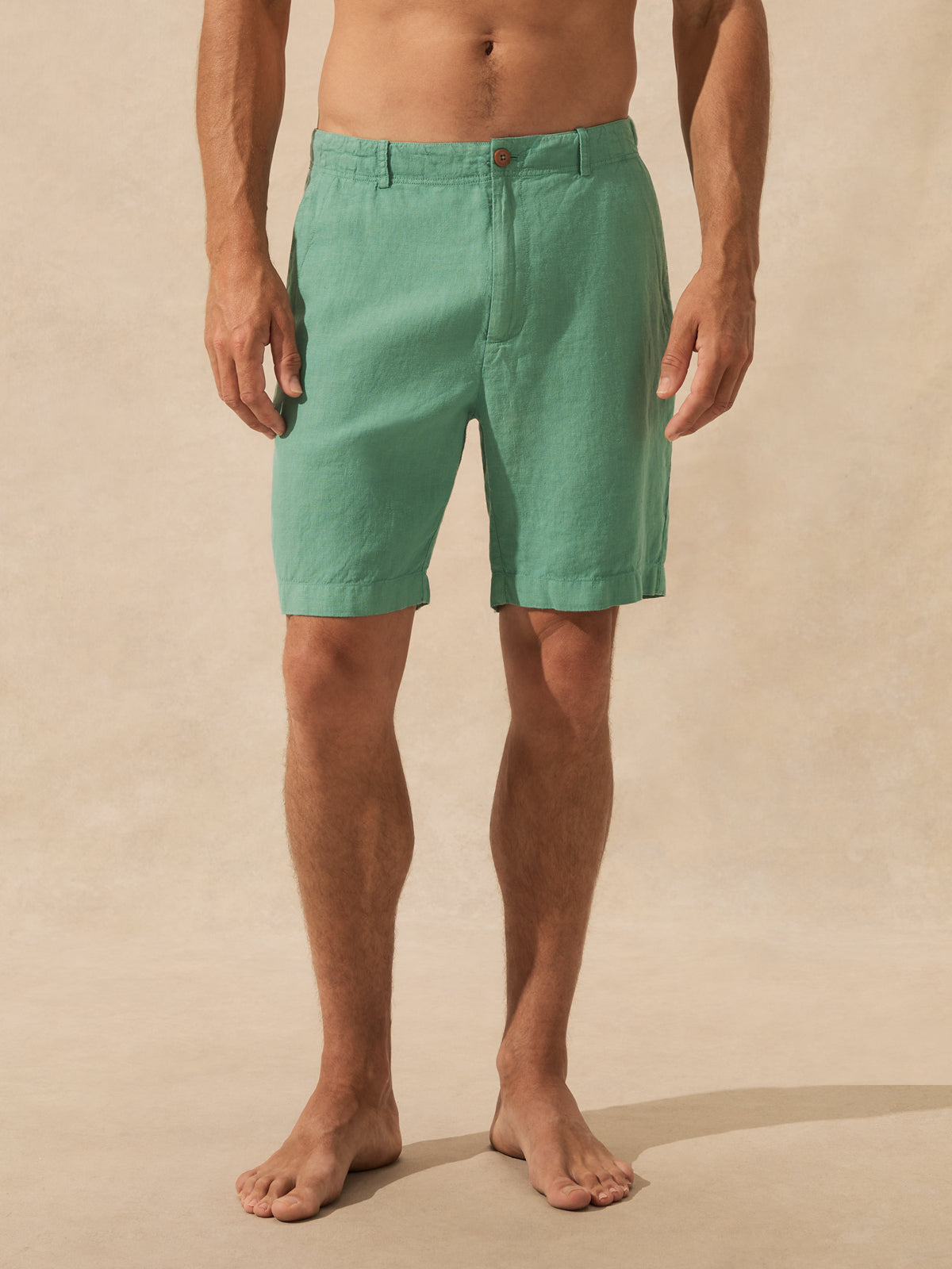 A man wearing green linen shorts, shown from the waist down against a warm beige textured studio backdrop.