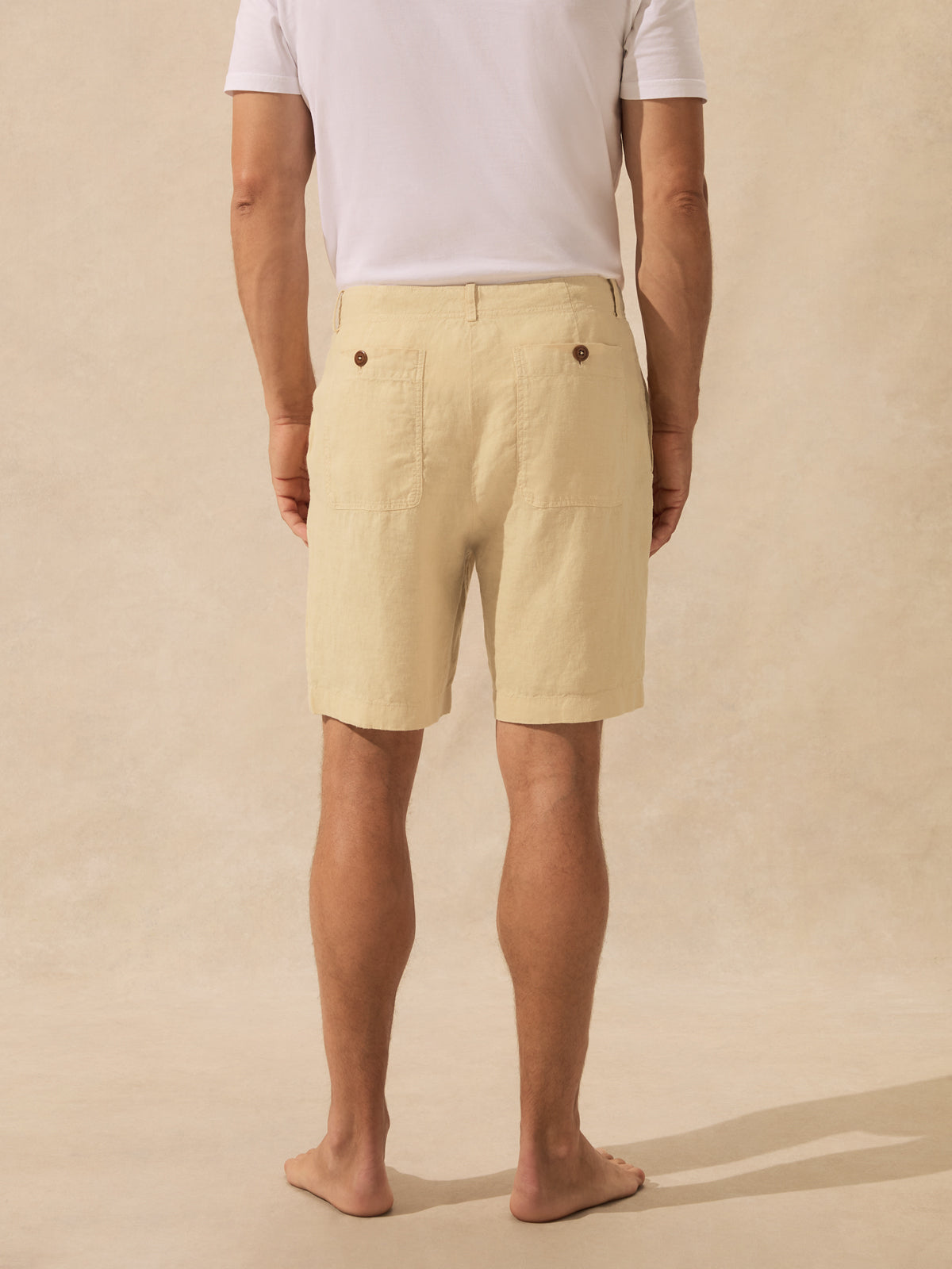 A man viewed from the back wearing light beige linen shorts and a white t-shirt, standing barefoot against a textured beige backdrop.