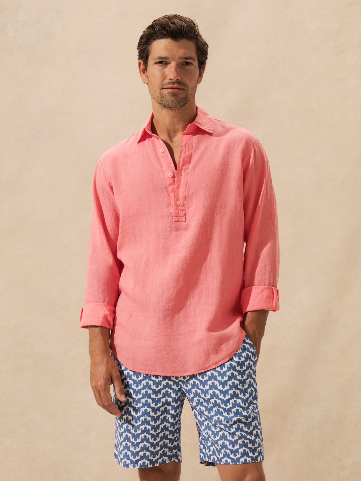 A man wearing a coral pink linen henley shirt and blue patterned shorts, standing against a textured beige background.