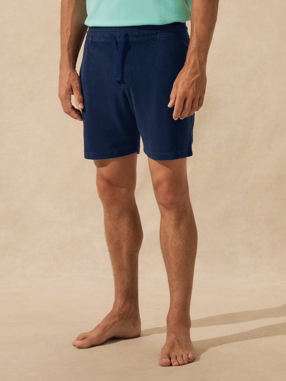 A man wearing navy blue drawstring shorts and a mint green top, standing barefoot against a warm beige textured studio background.