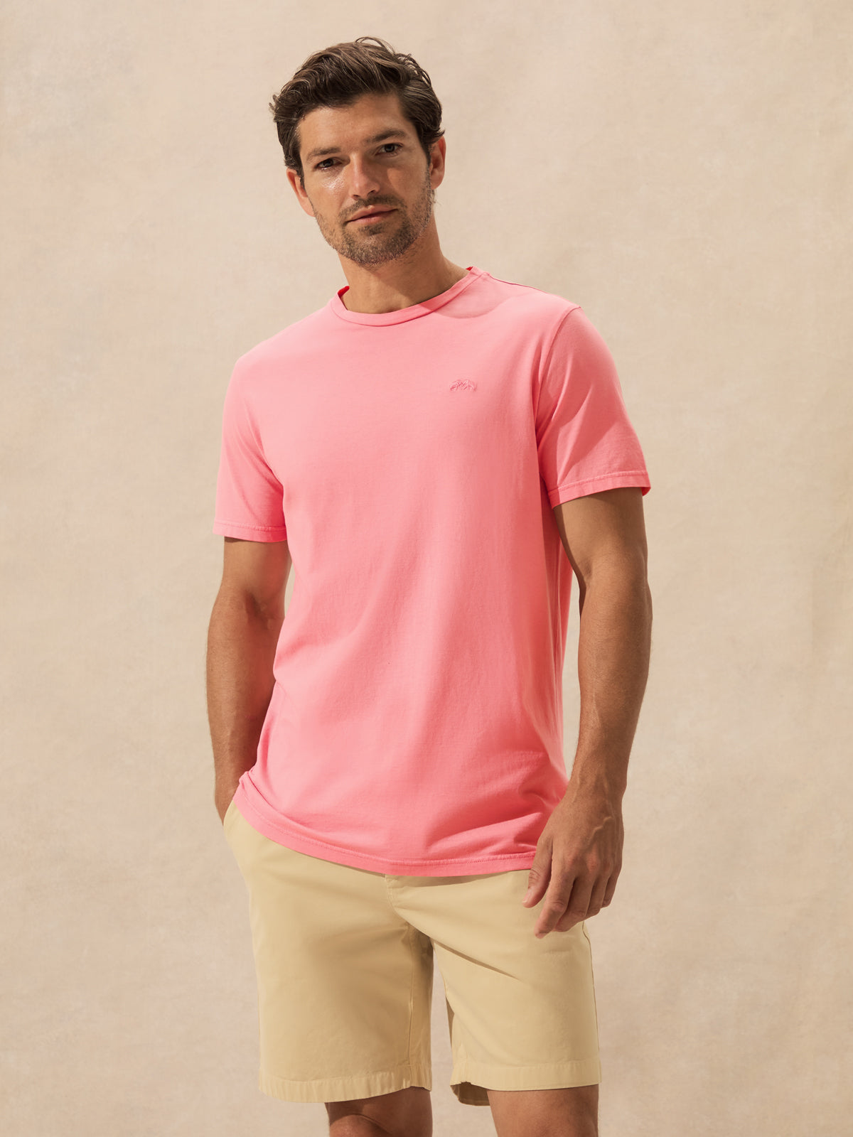 A man wearing a bright pink short-sleeve crewneck t-shirt and beige shorts, standing against a textured beige studio backdrop.