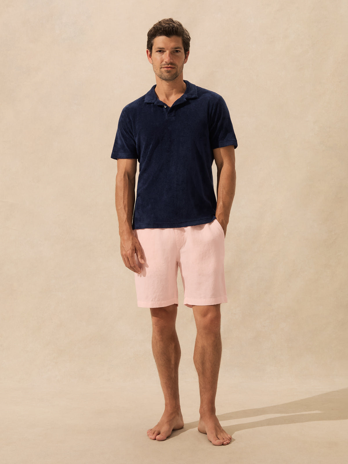 A man stands barefoot against a warm beige textured backdrop, wearing a short-sleeve navy polo shirt and light pink shorts.