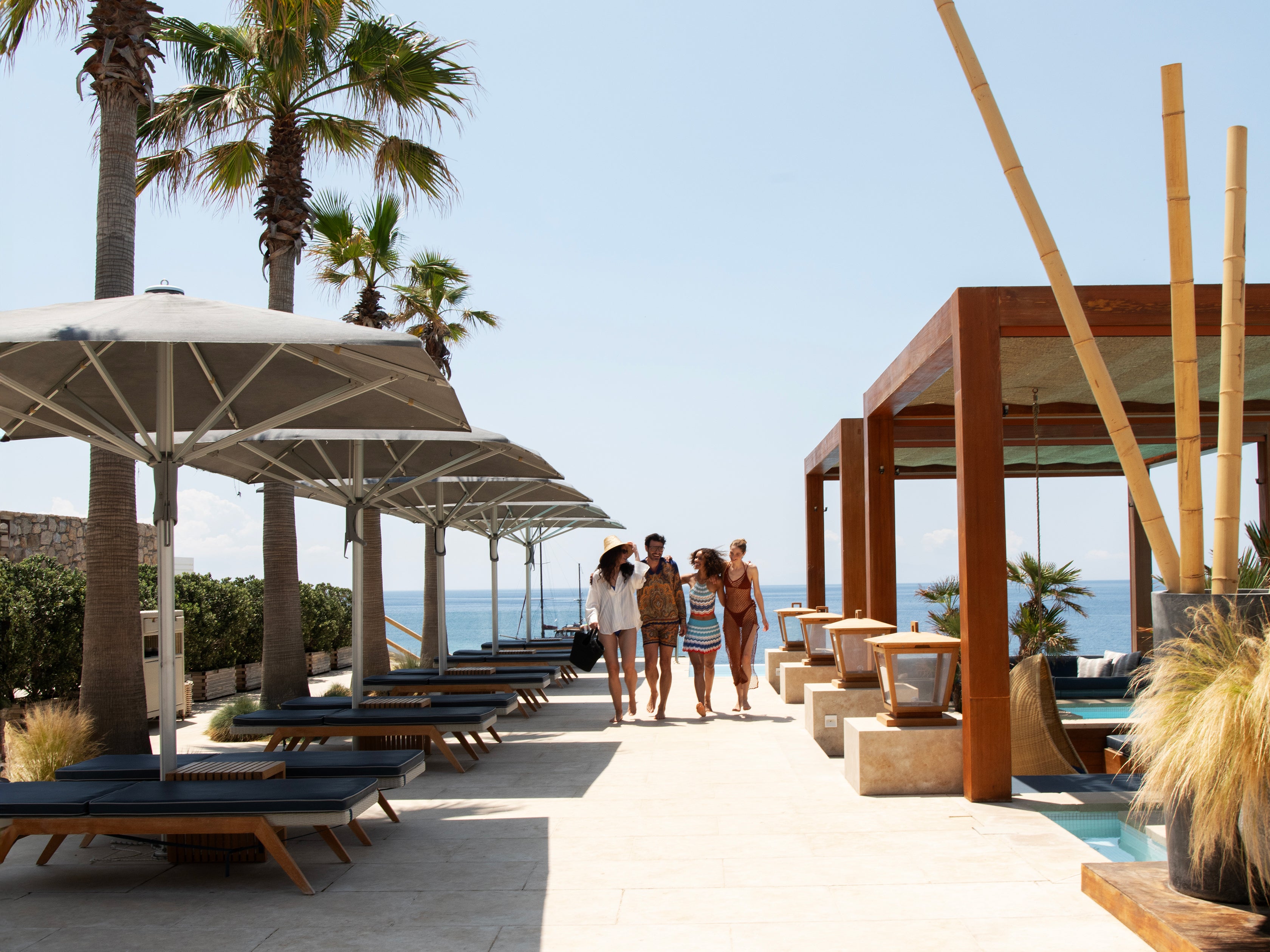 People walking along a sunlit outdoor area with palm trees and ocean view.