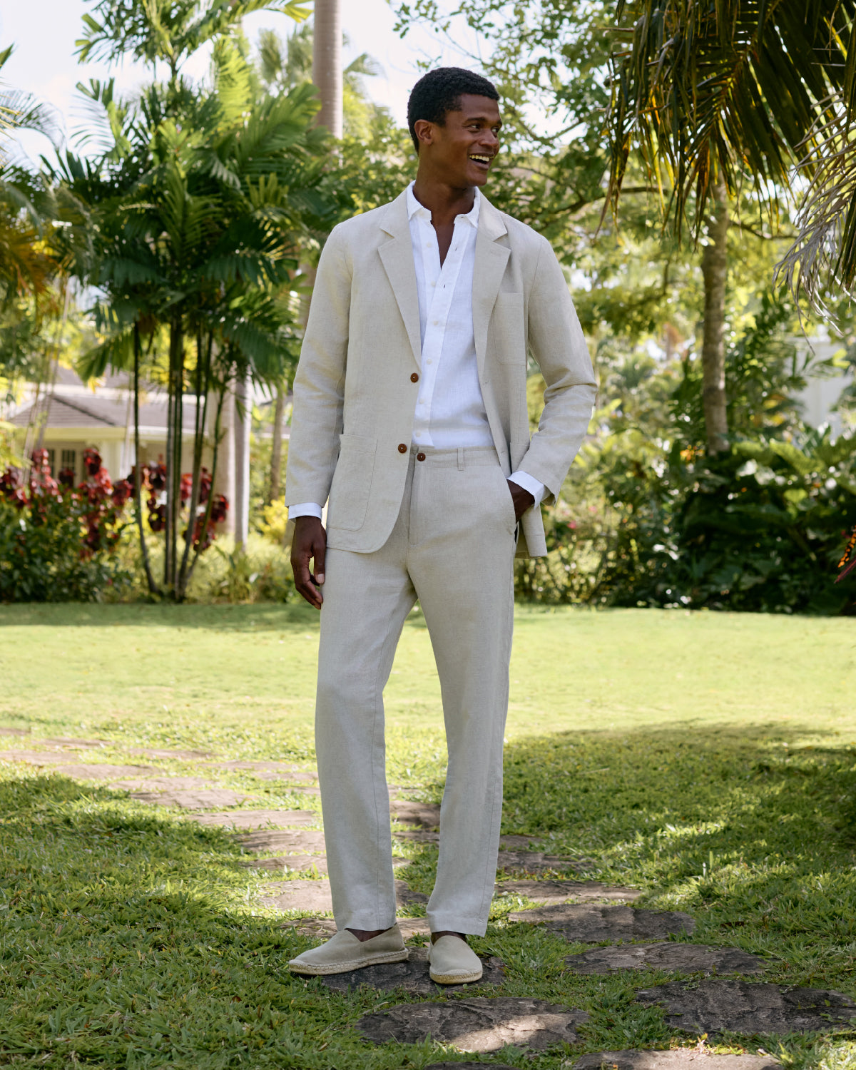 Man in a beige linen jacket, white linen shirt and beige pants standing on a stone path in a garden.