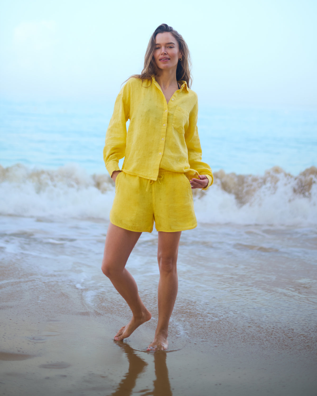 Woman in a yellow linen outfit standing on a beach with waves in the background