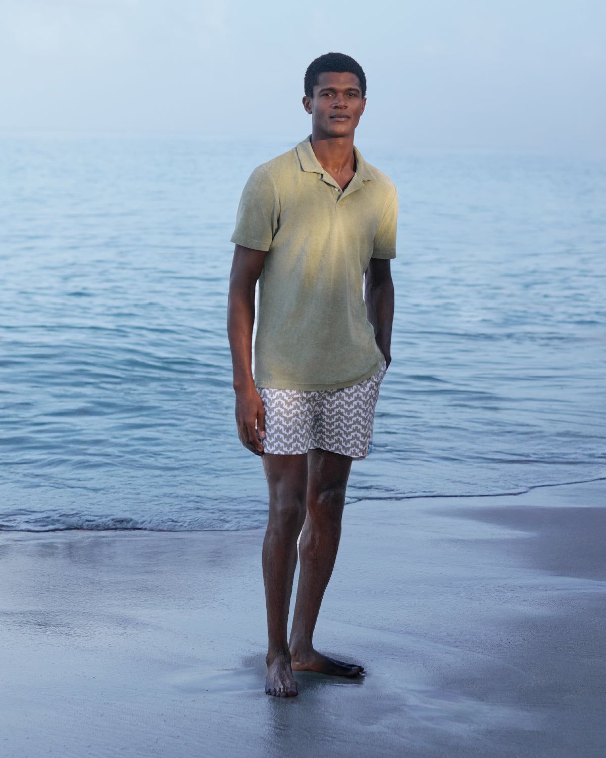 Man standing on a beach wearing a light green polo shirt and patterned swim trunks.