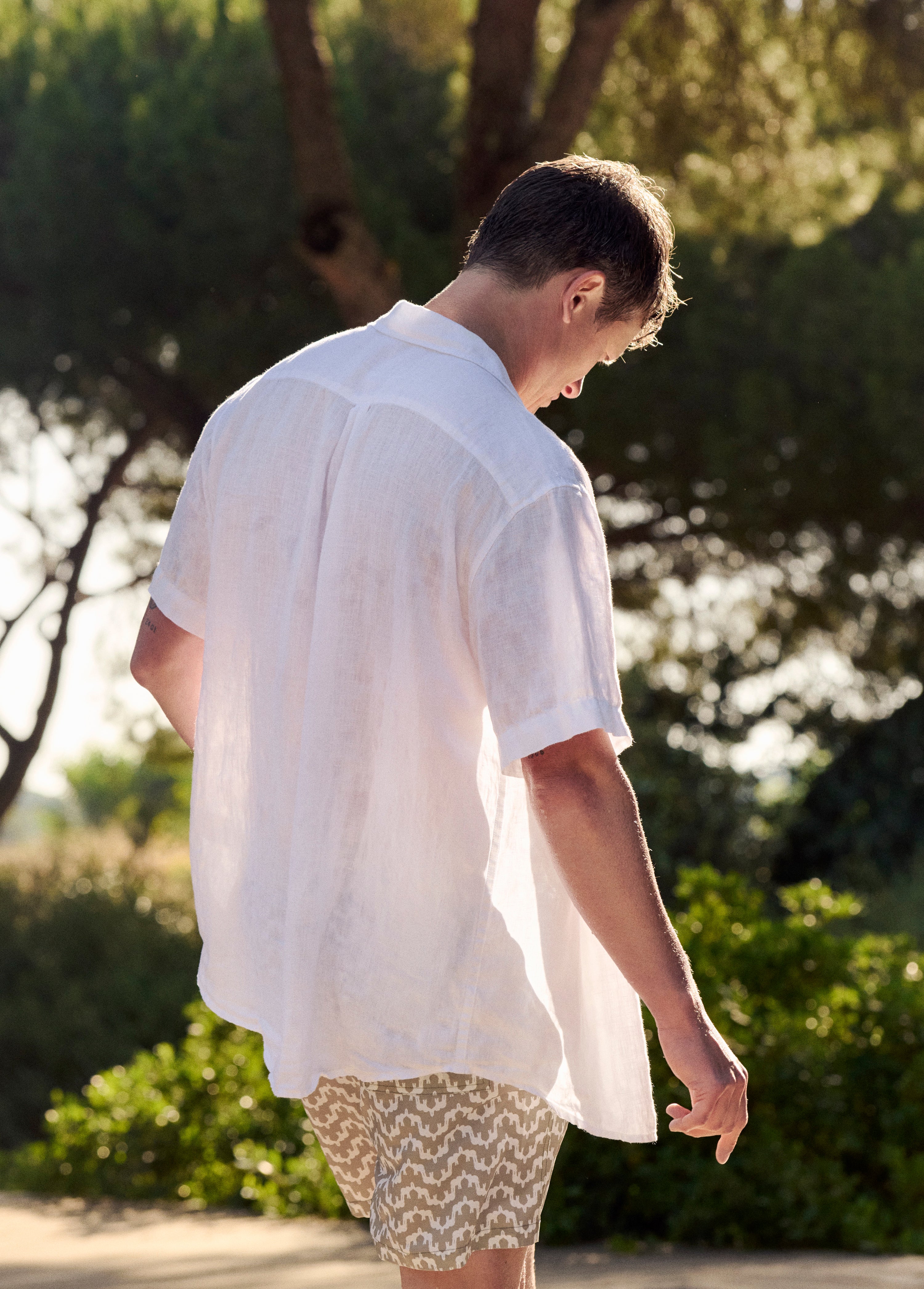 Actor Ed Speleers wearing a white short-sleeve button-up shirt and beige patterned shorts, photographed from behind in an outdoor park setting with trees and sunlight.