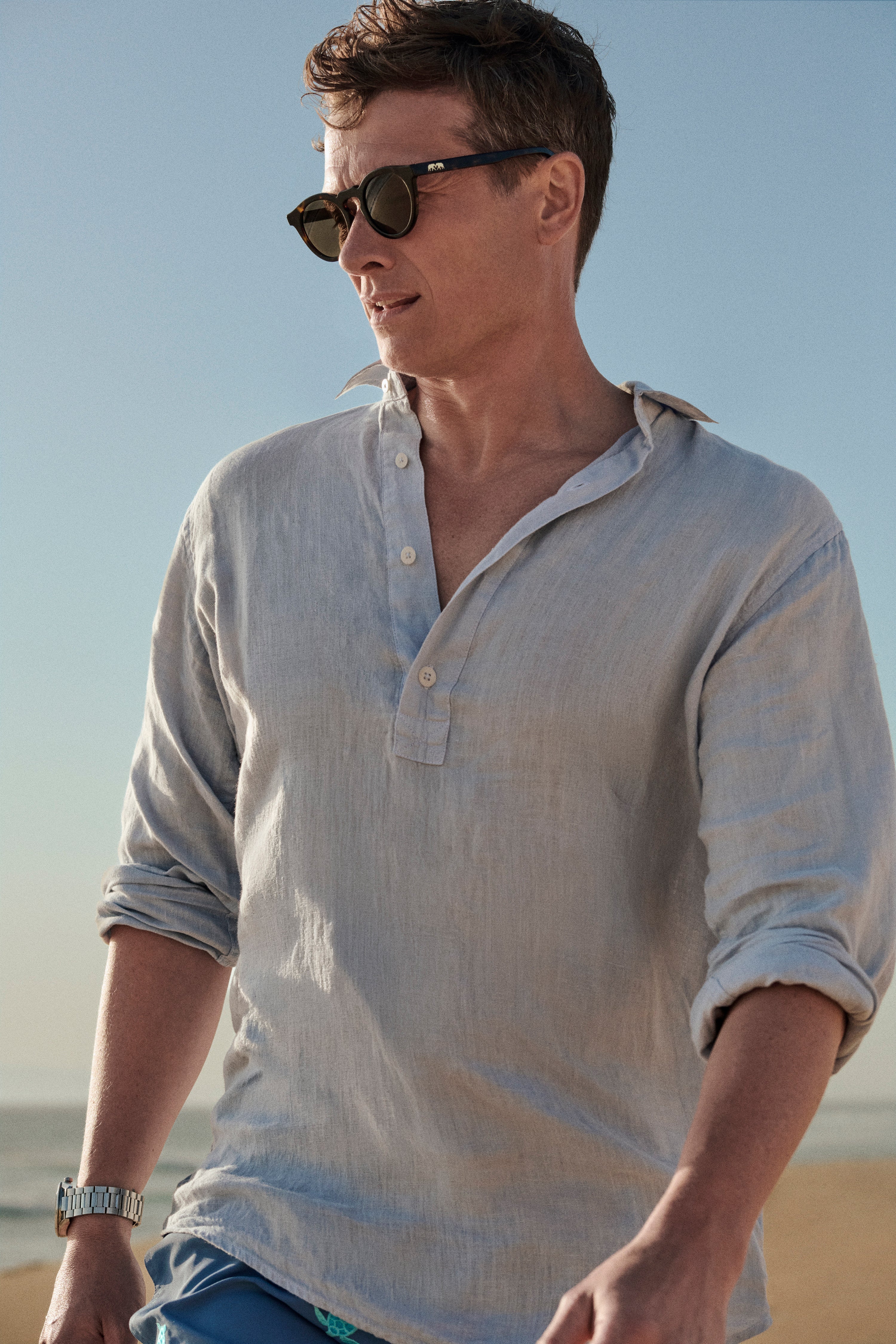 Actor Ed Speleers wearing a light blue linen shirt and dark sunglasses, standing outdoors against a clear blue sky.