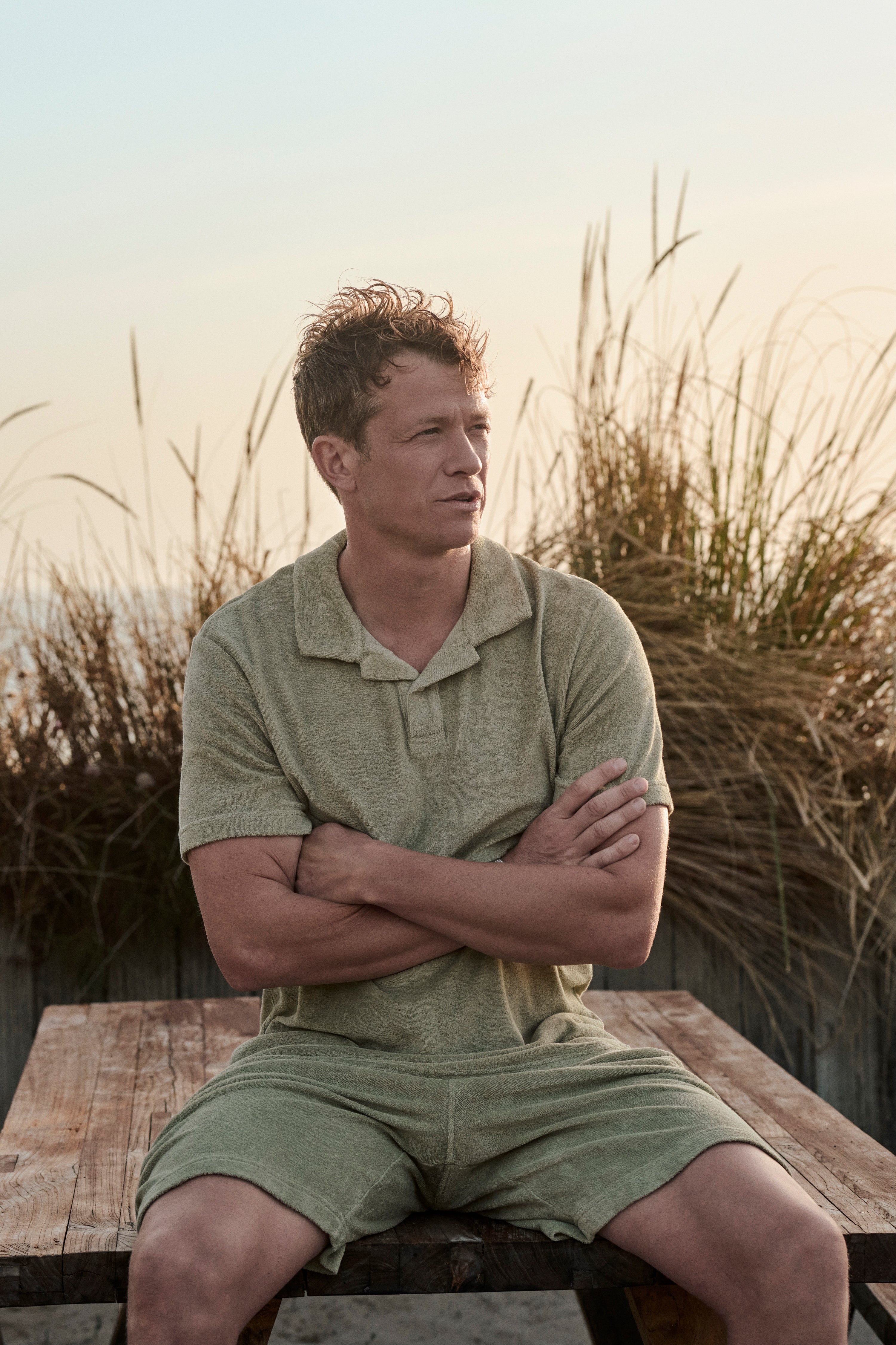 Ed Speleers sitting on a wooden bench outdoors at sunset, wearing a muted sage green short-sleeve polo shirt and matching shorts, with tall grasses in the background.