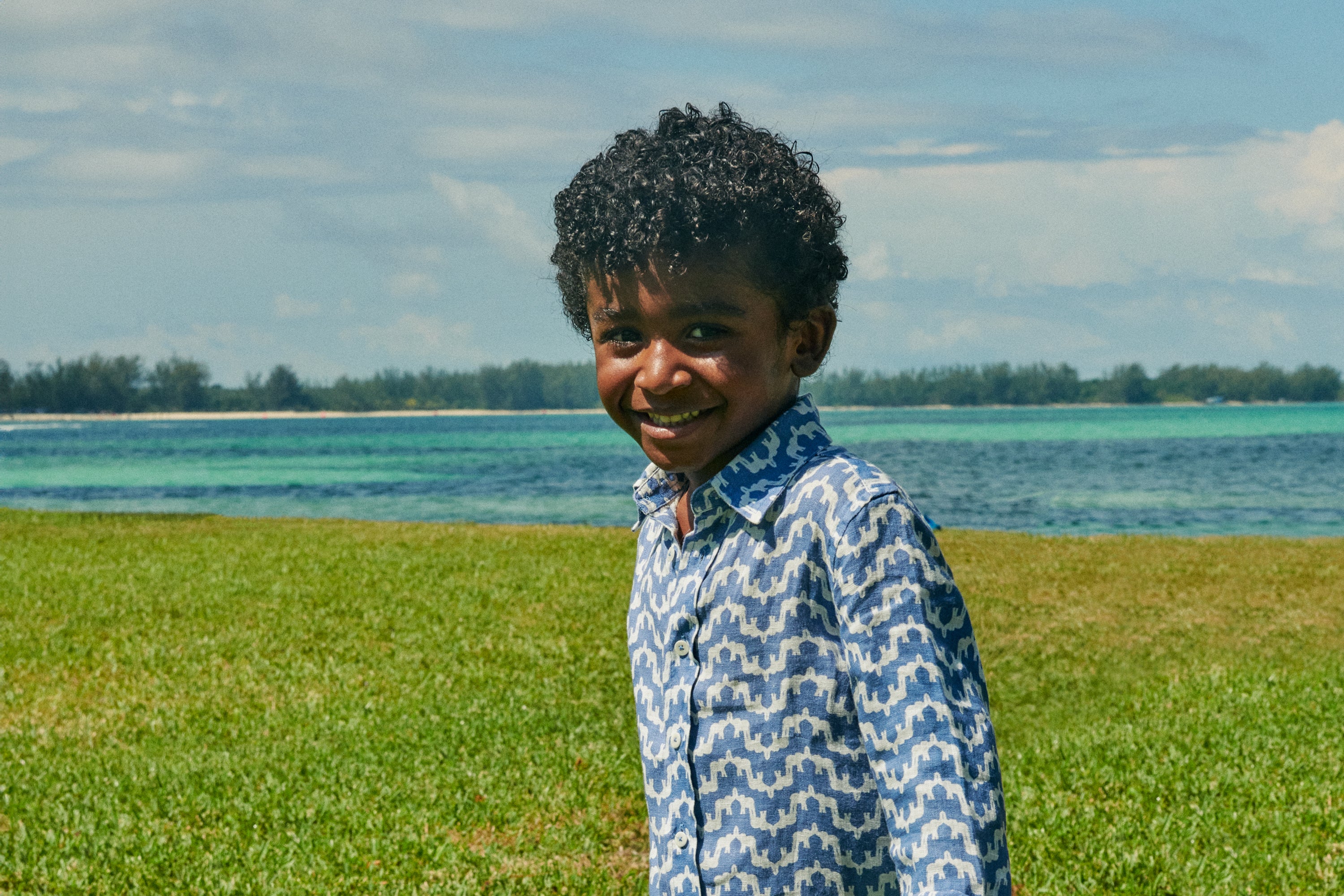 Child standing on a grassy field with a body of water and trees in the background wearing a blue patterned linen shirt.