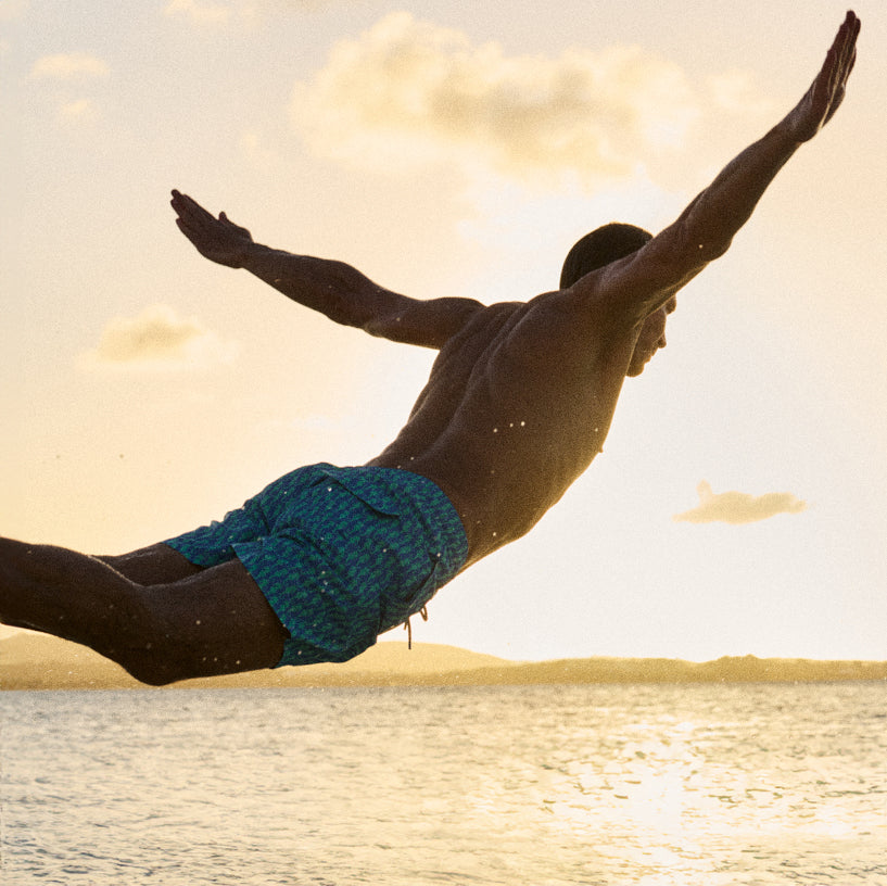 A man diving horizontally into the ocean at sunset, wearing blue patterned swim trunks. The scene is backlit by the warm golden glow of the setting sun, with clouds and water visible in the background.