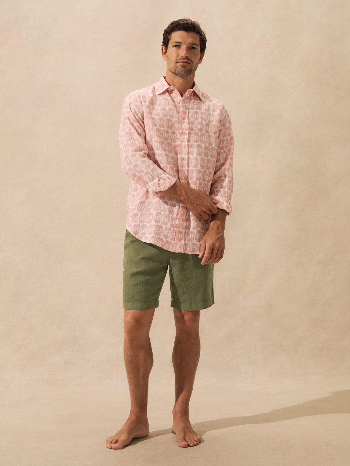 A man stands barefoot against a warm beige textured backdrop, wearing a light pink long-sleeve button-up shirt with a white geometric pattern and olive green shorts.