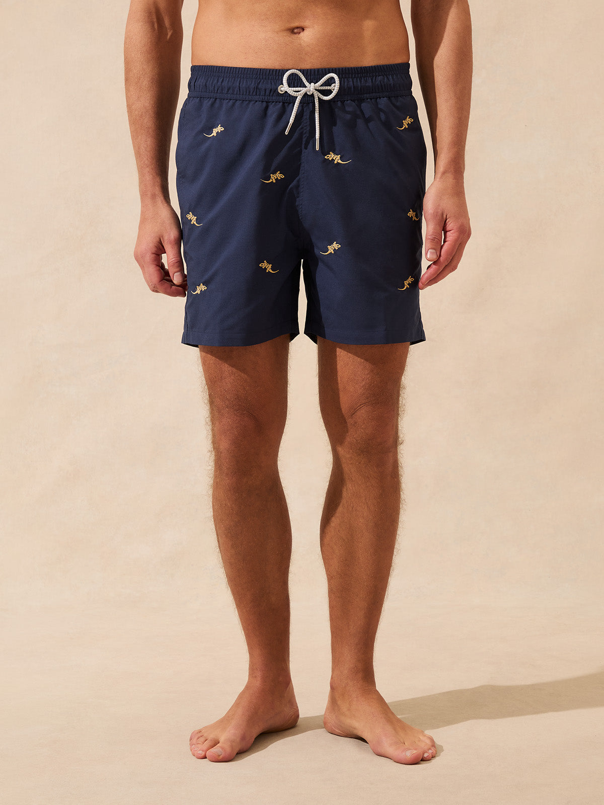Man wearing navy swim shorts with embroidered geckos on a beige background