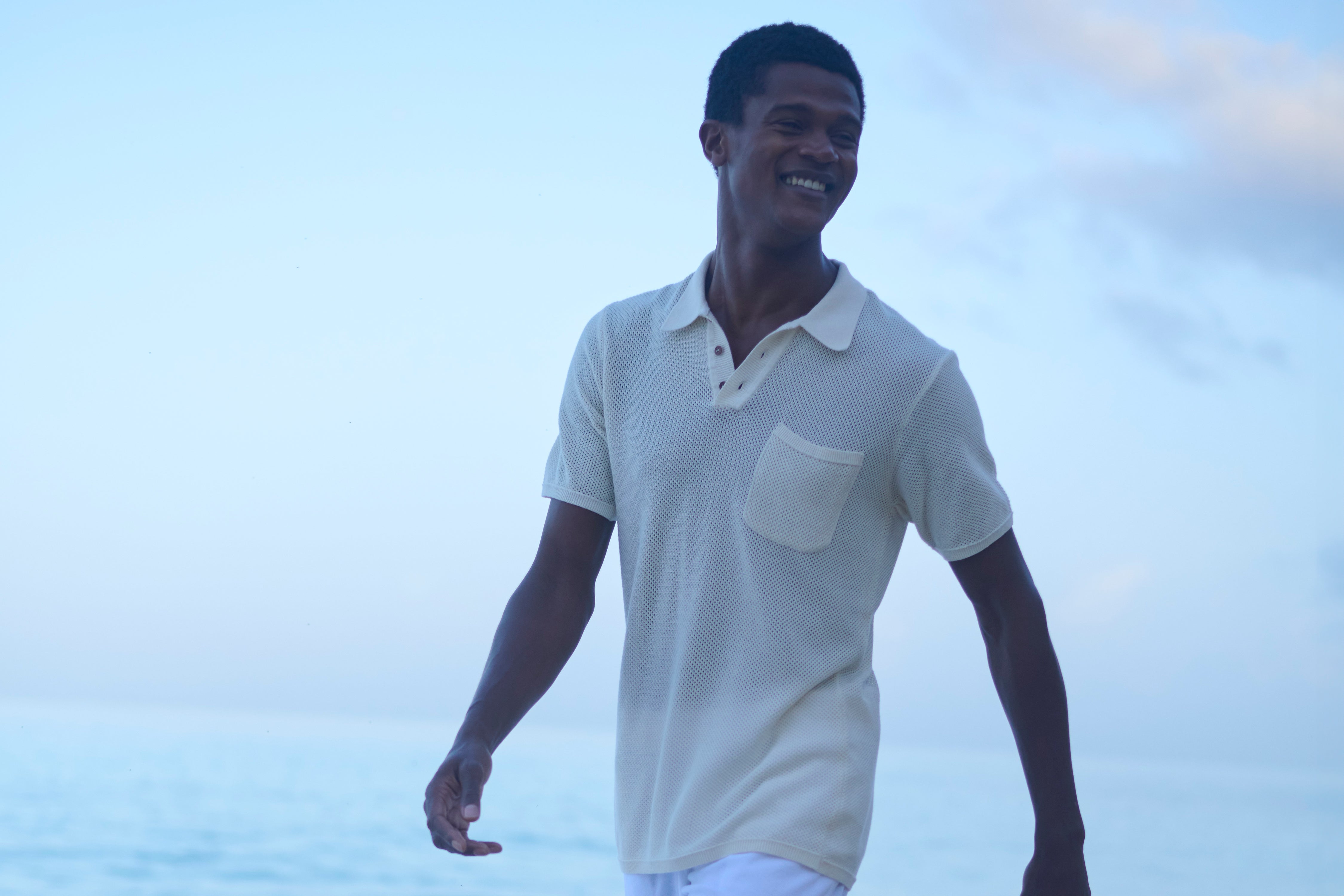 Man wearing a cream knitted polo shirt with a pocket, standing against a light blue sky.