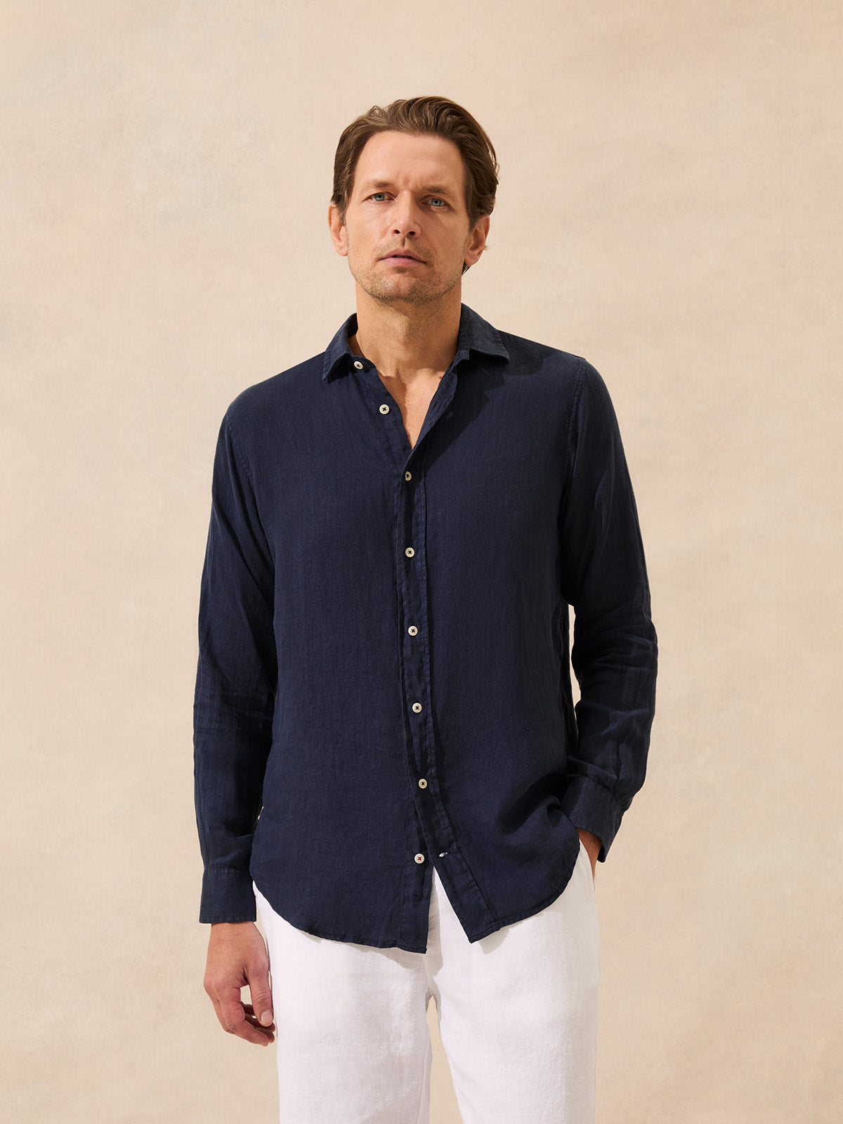 Man wearing a navy linen button-up shirt and white pants against a beige background