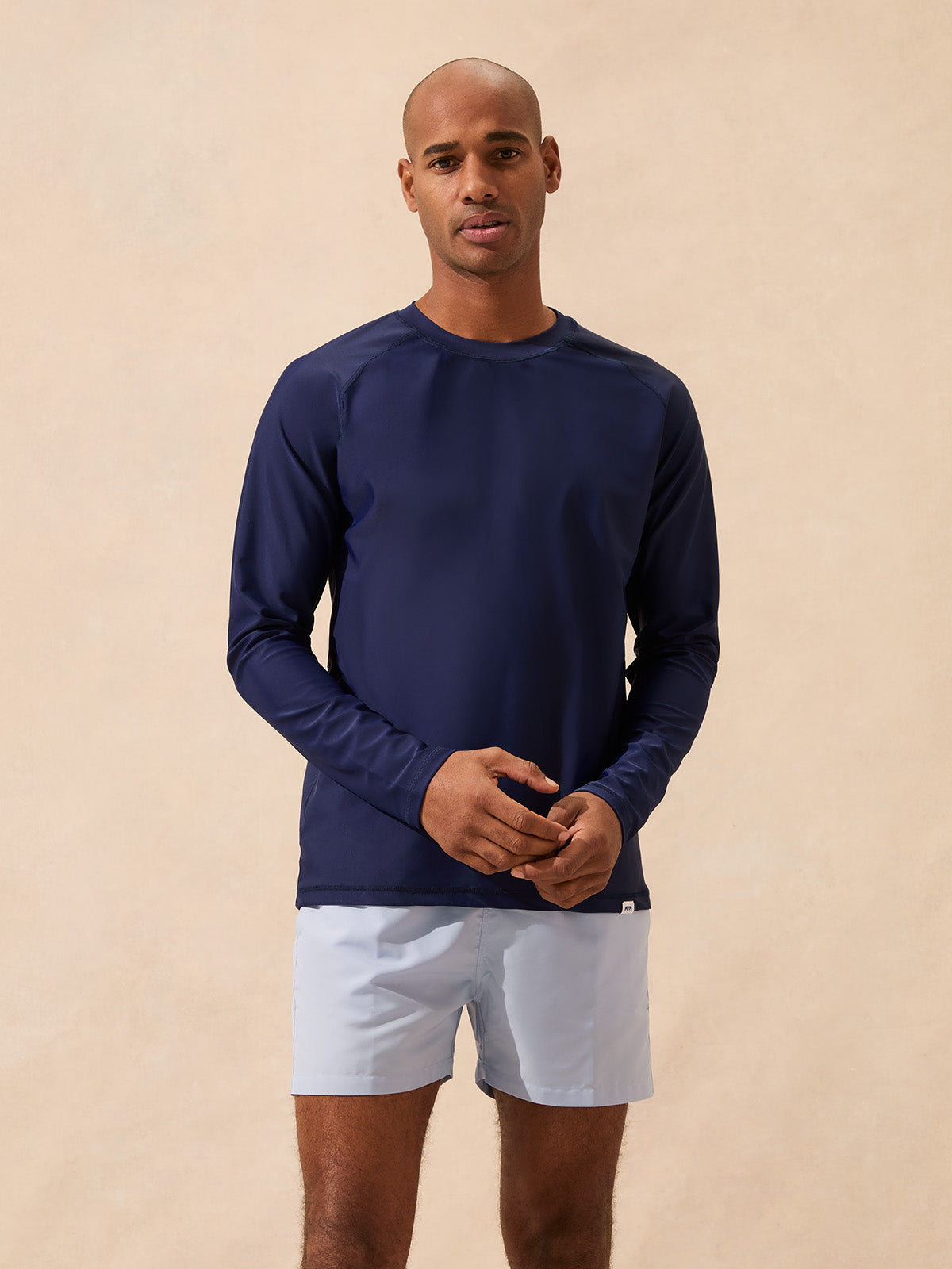 Man wearing a navy long-sleeve rash vest and light blue swim shorts against a beige background