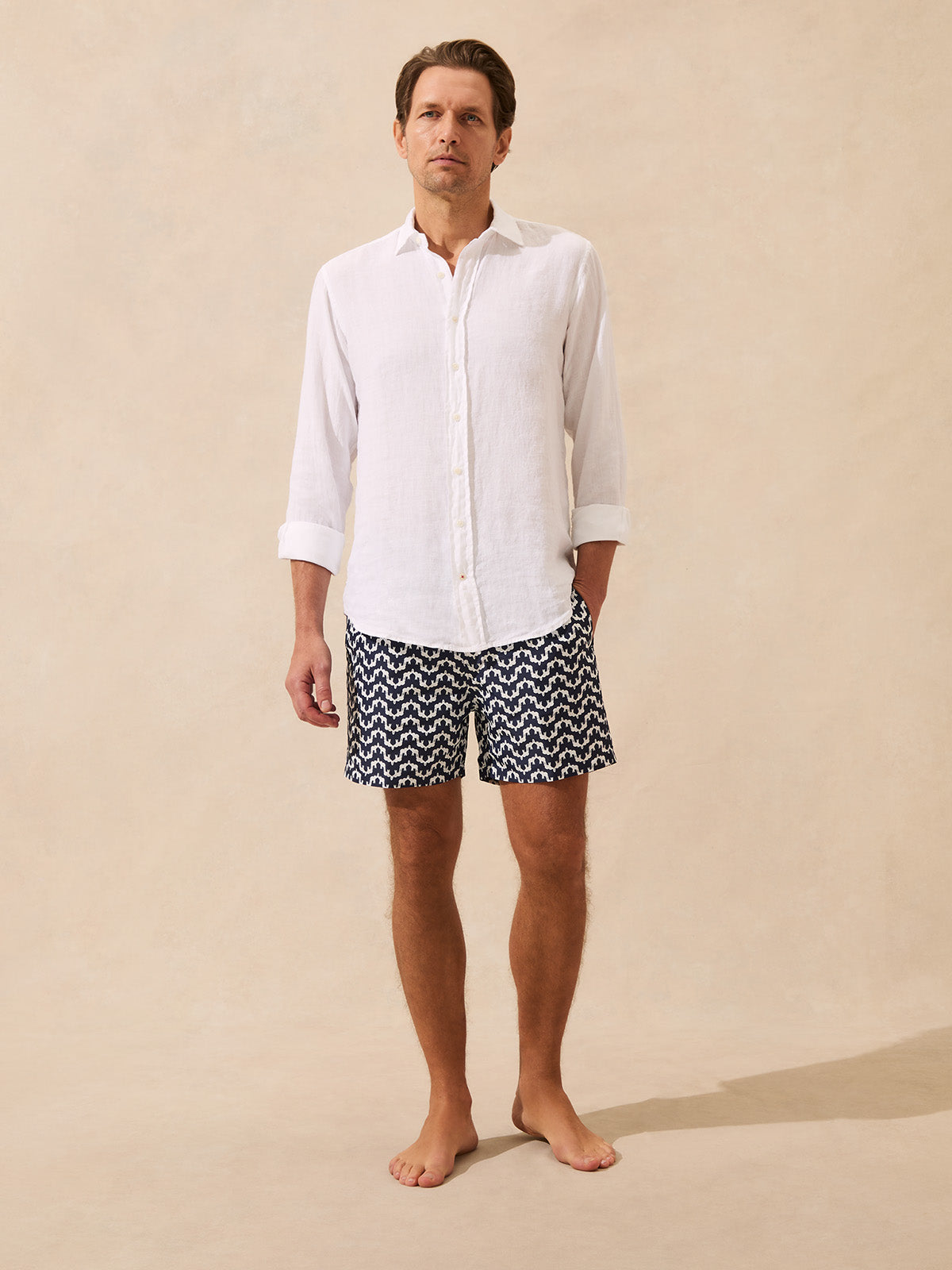 Man wearing a white linen shirt and patterned swim shorts on a beige background