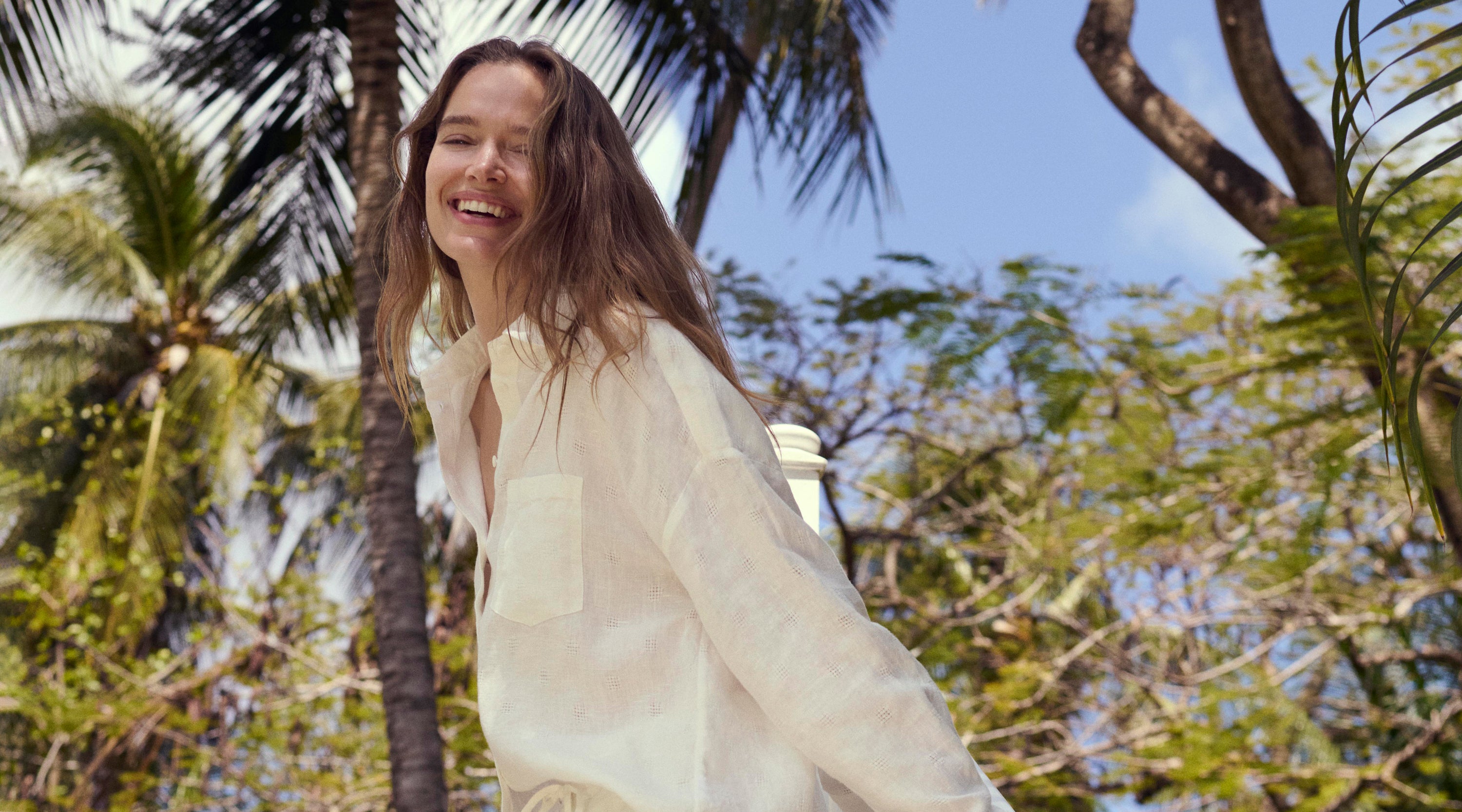 Woman in a white shirt standing among palm trees with a clear blue sky.