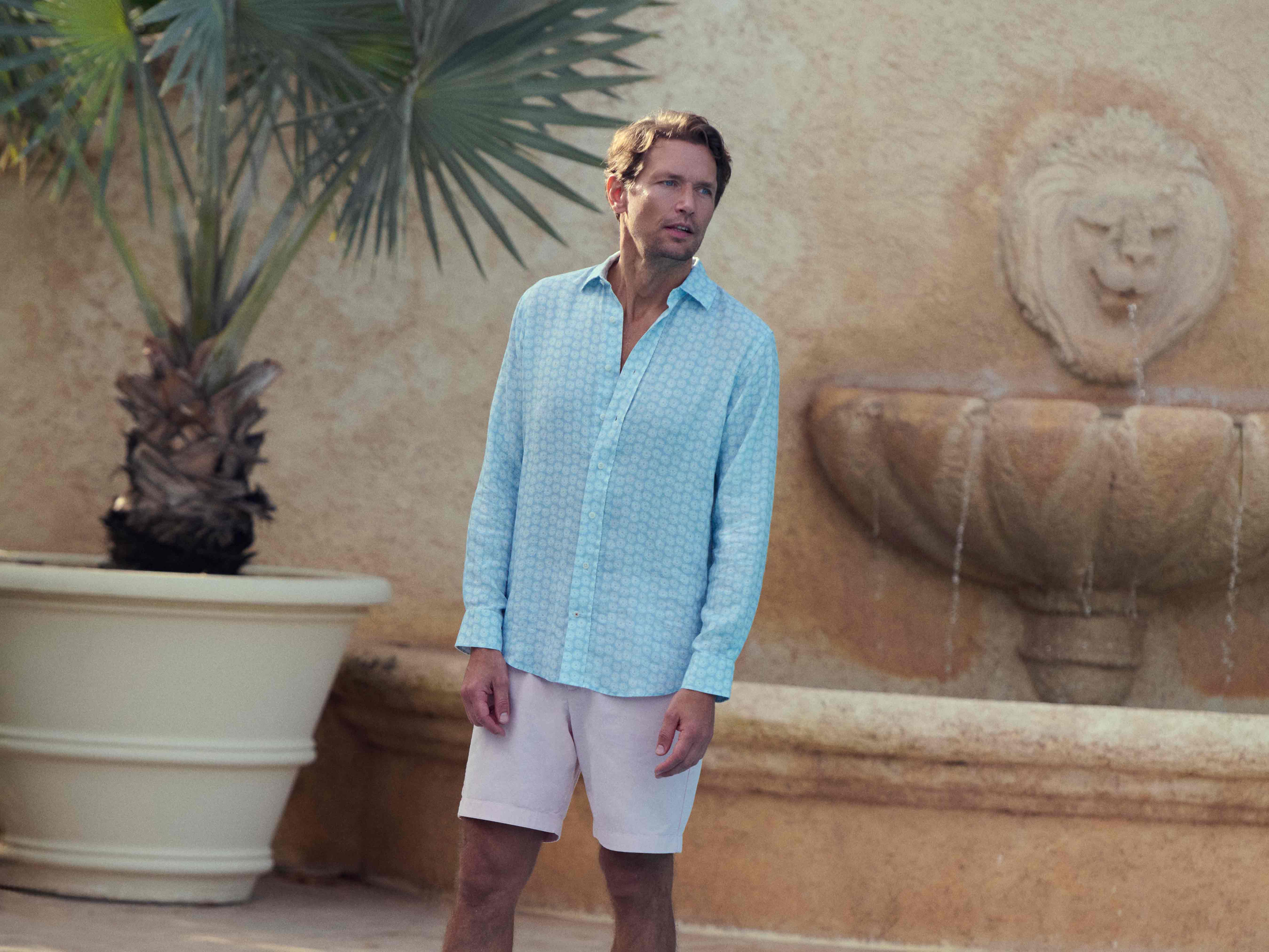 Man in light blue shirt and white shorts standing in front of a stone fountain with palm tree in the background